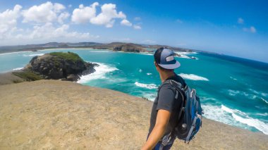A young man in a full cap walking on a cliff of Tanjung Aan Beach on Lombok, Indonesia and taking a selfie. Sea water is calm, shimmering with many shades of blue. Solo traveller in an unspoiled area