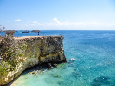 A man in shorts walking at the edge of cliffs of Pink Beach in Lombook, Indonesia. The man enjoys the view. The sea water has clear turquoise colour. Happiness and adventurous travelling