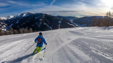 A man with a small backpack skiing on a perfeclly groomed slopes in Bad Kleinkirchheim, Austria. There are lots of snow caped mountains around him. Few trees on the side. Winter sports.