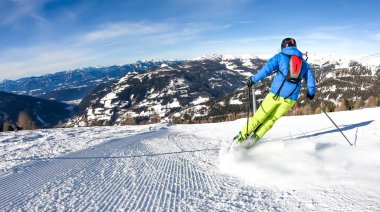 A man with a small backpack skiing on a perfectly groomed slopes in Bad Kleinkirchheim, Austria. There are lots of snow caped mountains around him. Few trees on the side. Winter sports.