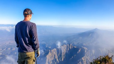 A young man standing of the top of volcano Inerie in Bajawa, Flores, Indonesia. He is enjoying the beautiful view on volcanic island. There are some clouds below him. Discovering while travelling.