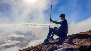 A young man sitting on the side of volcano Inerie in Bajawa, Flores, Indonesia. He is surrounded by clouds, as if she was walking on them. Clear blue sky. Natural phenomenon. He hold a walking stick