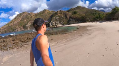 A young man walking on idyllic Koka Beach while taking a selfie. Hidden gem of Flores, Indonesia. There are tall hills overgrown with plants in front of him. Paradise like island. Island life.