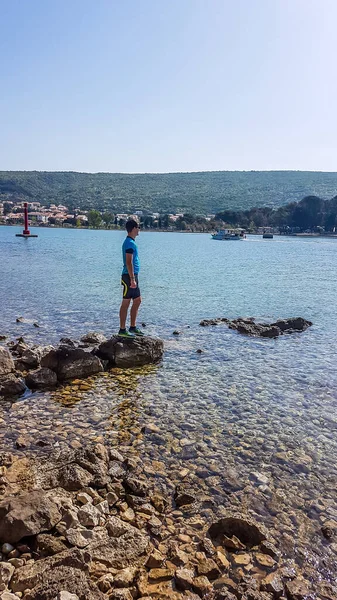 A young man in a biking outfit, standing on a rock in a shallow sea water and admiring the view in front of him. Water has many shades of blue. Sea is very calm. Clear and sunny day.