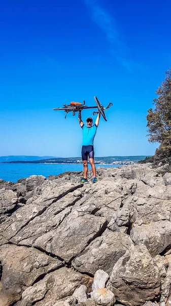 A young man in a sporty outfit standing on a rocky shore of Mediterranean Sea and rising his bike above his head in a gesture of triumph and victory. Sharp rock shaped by the water. Clear day.