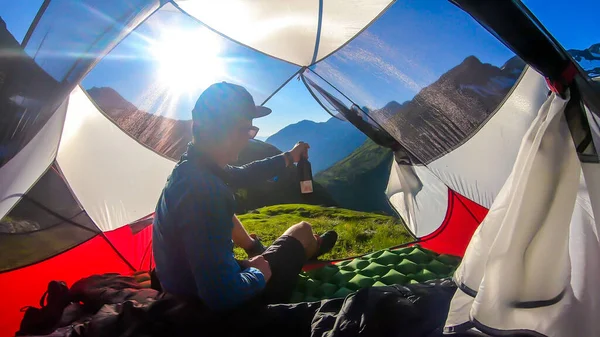 A man camping in a wilderness. He sits in a small tent, placed on a top of a mountain peak, waiting for the sunset with a bottle of wine in his hand. High mountains around. Spring in alpine valleys.