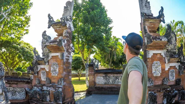 A young man taking a selfie under the entrance to Hindu temple in Lombok, Indonesia. The gate has vivid orange color with many decorations. Discovering new cultures and traditions.