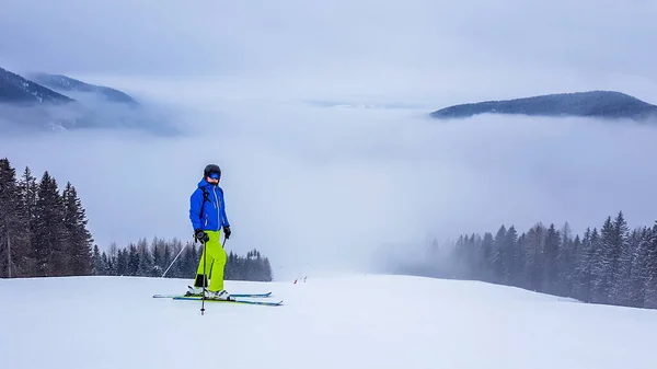 A skier in blue jacket and green trousers standing on a snowy slope with a very thick fog in the valley. The visibility is very low. Not favourable condition for skiing in Goldeck, Austria. Overcast.