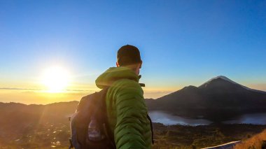 A man in a jacket standing on top of Mount Batur, Bali, Indonesia and waiting for the sunrise. There is Mount Rinjani in the back (Lombok) and Volcano Agung on the side. Fog in the valley. Mysterious
