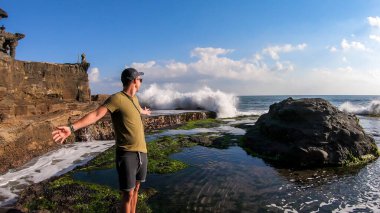 A man standing next to an edge of a cliffs, enjoying the waves splashing on cliffs, next to Tanah Lot Temple, Bali, Indonesia. Power of the nature. The man is enjoying his time and surrounding nature