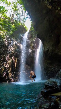 A man in swimsuit jumping from the slippery rocks in front of Gitgit Twin Waterfall, Bali, Indonesia. The waterfall is surrounded by rocks. The man enjoying the beauty of the nature. Adventure