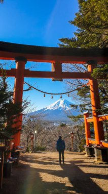 A man standing under the Torri gate, leading to Chureito Pagoda in Japan and Mt Fuji in the back, on a clear, wintery day. The top parts of the volcano are covered with a layer of snow. Holly mountain