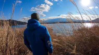 A man walking in between golden grass at the shore of Kawaguchiko Lake, Japan with the view on Mt Fuji. He shovels the grass sideways with his hands. The mountain surrounded by clouds. Adventure
