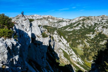 A man with a hiking backpack standing at the mountain ledge in Hochschwab region in Austria. There are many mountain ranges around her. The slopes overgrown with mountain pine, partially barren rocks