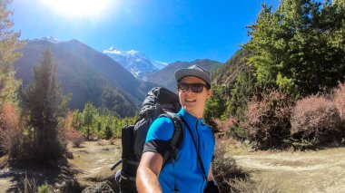 A man taking a selfie while trekking along Annapurna Circuit in Nepal. He is enjoying the view and trek. There is a lush green Himalayan valley around him. Snow caped mountains in the back