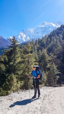 A man trekking along Annapurna Circuit in Nepal. He is enjoying the view and trek. There is a lush green Himalayan valley around him. Snow caped mountains in the back. Happiness and achievement.