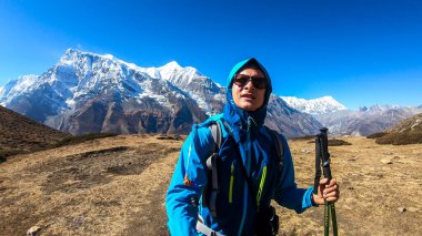 Man taking a selfie while trekking on the Annapurna Circuit Trek, Himalayas, Nepal. Snow caped Annapurna chain in the back. Lots of dried grass. High altitude, massive mountains. Freedom and adventure
