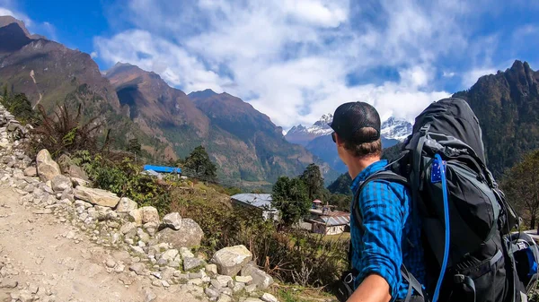 A man with a big black backpack hikes along a pathway on Annapurna Circuit Trek in Himalayas, Nepal, while taking a selfie. There are massive mountain chains around him. Exploration and adventure