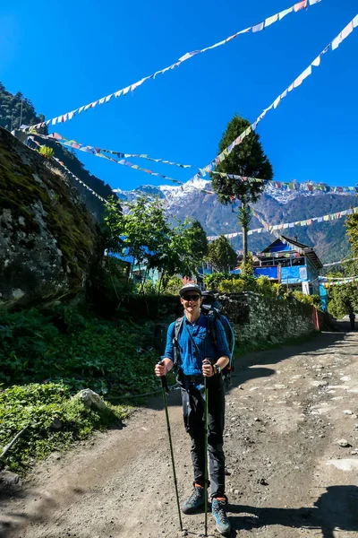 A man in hiking outfit standing in front of a small Himalayan village. Colorful prayer flags waving above the Himalayan peaks along Annapurna Circuit Trek, Nepal. Meditation and peace of mind.