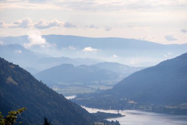 A panoramic view on the Lake Ossiacher located in between high Alpine peaks in Austria. The surface if the lake reflects golden sunbeams A bit of overcast. Few tree brunches entering the frame. Calm clipart