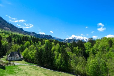 A a small chapel at the edge of the forest the panoramic view on Baeren Valley in Austrian Alps. The highest peaks are snow-capped. Lush green pasture. Clear and sunny day. High mountain chains. clipart