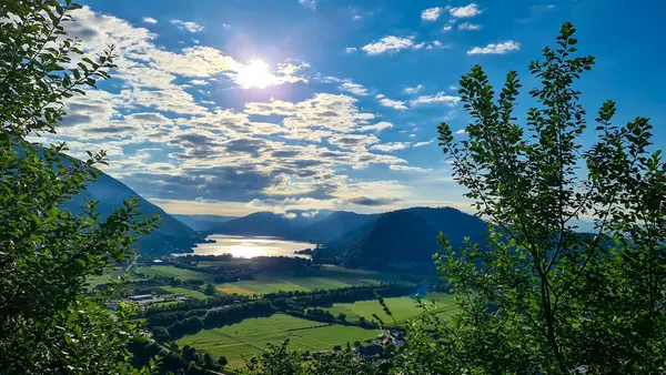 Avusturya, Carinthia 'daki yüksek Alp tepelerinde gömülü Ossiach Gölü manzarası. Göl altın güneş ışınlarını yansıtırsa yüzey. Çerçeveye birkaç ağaç brançı giriyor. Oswaldiberg