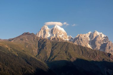 First sunrise reaching the peaks of Ushba in Caucasus, Georgia. Cloudless sky above the high and snow-capped mountains. The hills below are shrouded with shadows. Daybreak. High and desolate mountains clipart