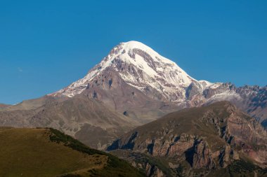 Kafkasya, Georgia 'daki Kazbeg Dağı' nın bulutsuz manzarası. Orada kar kaplı tepenin ve Gergeti Buzulu 'nun altında çorak ve taşlı yamaçlar var. Sükunet. Doğal bir ilaç. Devasa buzul ayağı.