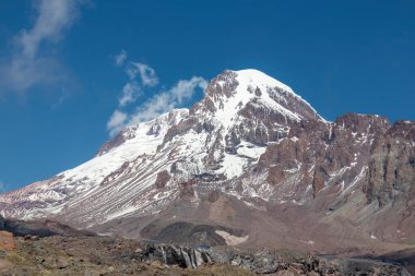 Kafkasya, Georgia 'daki Kazbeg Dağı' nın bulutsuz manzarası. Orada kar kaplı tepenin ve Gergeti Buzulu 'nun altında çorak ve taşlı yamaçlar var. Sükunet. Doğal bir ilaç. Devasa buzul ayağı.