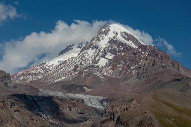 Kafkasya, Georgia 'daki Kazbeg Dağı' nın bulutsuz manzarası. Orada kar kaplı tepenin ve Gergeti Buzulu 'nun altında çorak ve taşlı yamaçlar var. Sükunet. Doğal bir ilaç. Devasa buzul ayağı.