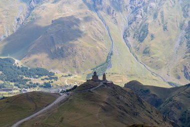Gürcistan 'ın Stepansminda kentindeki Gergeti Trinity Kilisesi' ne uzaktan bakış. Kilise yüksek bir Kafkas dağında yer almaktadır. Kilisenin üstündeki açık ve mavi gökyüzü. Arkada yüksek dağ zincirleri var..