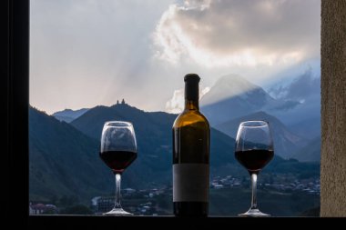 Georgian red wine with a panoramic evening on Mount Kazbegi in the Greater Caucasus Mountains in Georgia. Clouds are covering the snow-capped summit. Sunset, sunrise. Romantic date, two wine glasses. clipart