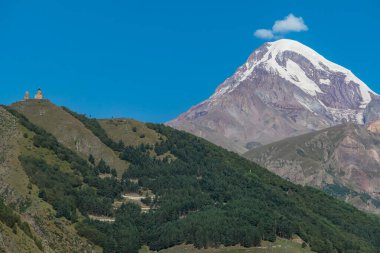 Gürcistan, Stepansminda 'daki Gergeti Trinity Kilisesi' ne uzaktan bakış. Kilise, Büyük Kafkas Dağları 'nda yer almaktadır. Arkadaki Kazbegi Dağı 'nın üstündeki açık gökyüzü. Yürüyüş yolu
