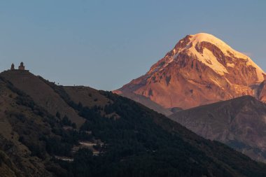 Gürcistan, Stepansminda 'daki Gergeti Trinity Kilisesi' ne uzaktan bakış. Kilise, Büyük Kafkas Dağları 'nda yer almaktadır. Arkadaki Kazbegi Dağı 'nın üstündeki açık gökyüzü. Gün doğumu, gün batımı