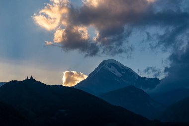 Gürcistan, Stepansminda 'daki Gergeti Trinity Kilisesi' ne uzaktan bakış. Kilise Büyük Kafkas Dağları 'nda. Bulutlar arkadaki kar kaplı Kazbegi Dağı' nı örtüyor. Gün doğumu, gün batımı.