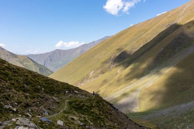 A female backpacker on a hiking trail with a panoramic view on the green hills and sharp ridges of the mountain peaks in the Greater Caucasus Mountain Range in Georgia, Kazbegi Region. Wanderlust. clipart