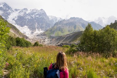 Ushguli 'nin Svaneti Bölgesi, Gürcistan' daki Büyük Kafkasya Dağları 'ndaki Shkhara Buzulu' nun panoramik manzarasını seyreden bir kadın. Gezgin şehveti, Alp otlağı. Çiçek açan.