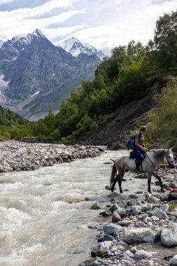 Adishischala nehrini bir atla geçen kadın. Gürcistan, Svaneti Bölgesi 'ndeki Büyük Kafkasya Dağları' ndaki Adishi Buzulu ve Tetnuldi, Gistola ve Lakutsia 'nın karlı zirvelerine bakın..