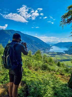 A man taking pictures at a viewing point near the top of Oswaldiberg with the view on the Lake Ossiach in Austrian Alps, Carinthia. The photographer enjoys a panoramic view on the Ossiacher Tauern. clipart