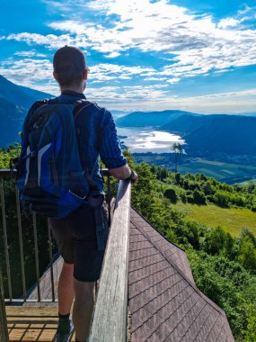 A man with a backpack standing at the viewing platform of a chapel at the top of Oswaldiberg with the view on the Lake Ossiach in Austrian Alps, Carinthia. The hiker enjoys panoramic view. Freedom clipart