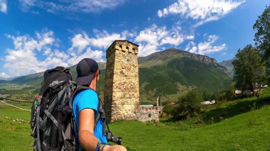 A man taking a picture wirh a lookout tower on a lush green pasture in Caucasus, Georgia. There are high mountain chains around. Clear and bright day with few clouds. Defense building. Hiking remedy clipart