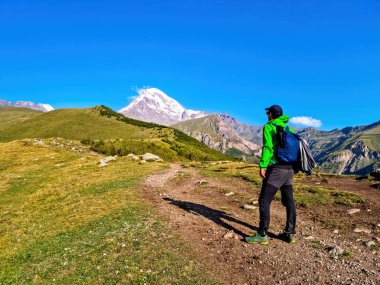 Kafkasya, Georgia 'daki Kazbeg Dağı' nın bulutsuz manzarasının tadını çıkaran bir adam. Buzulun karlı zirvesinin altında yemyeşil otlaklar ve yeşil tepeler var. Huzur ve sükunet. Doğa