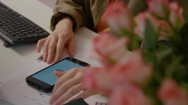 Kadın akıllı telefonuna odaklanmış, internetten alışveriş yapıyor. Ödemelerini sorunsuz yaparken mali tabloları ve grafikleri yakınlarda..