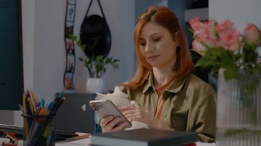Kadın ev ofisinde dinlenmekten zevk alıyor, beyaz kedisini nazikçe okşarken telefonunu kontrol ediyor. Çiçekler çevreye biraz renk katar.