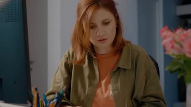 Şaşırmış bir kadın, evindeki evraklarla çevriliyken bilgisayarında bilgi arıyor. O, işinden sıkılmış ve memnun görünmüyor..