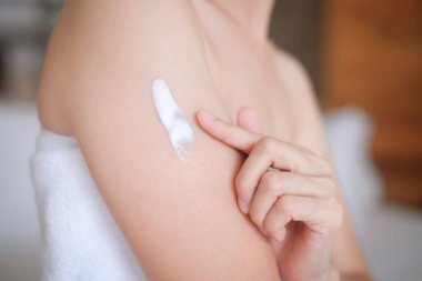 Asyalı kadın yatakta oturuyor ve koluna krem sürüyor.