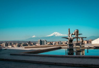Sabah konsept fotoğrafında dağ manzaralı şehir manzarası. Erivan, Ermenistan 'ın başkenti, Ararat Dağı' nın önünde. Modern sanat, havuzda üç sporcunun paslanmaz çelik heykelleri. Yüksek