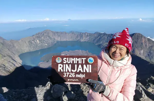 stock image Asian middle-aged woman is taking a photo at the top of Mount Rinjani while holding a summit marker placard with a clear blue sky background.