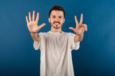 Genç adam kendine güvenen beyaz tişört giyiyor mavi arka planda soyutlanmış poz veriyor ve 8 numaralı parmaklarıyla gülümseyerek işaret ediyor..