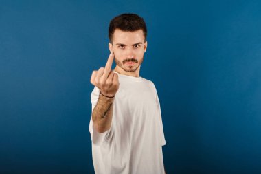 Handsome caucasian man wearing white t-shirt posing isolated over blue background showing middle finger doing fuck you bad expression.